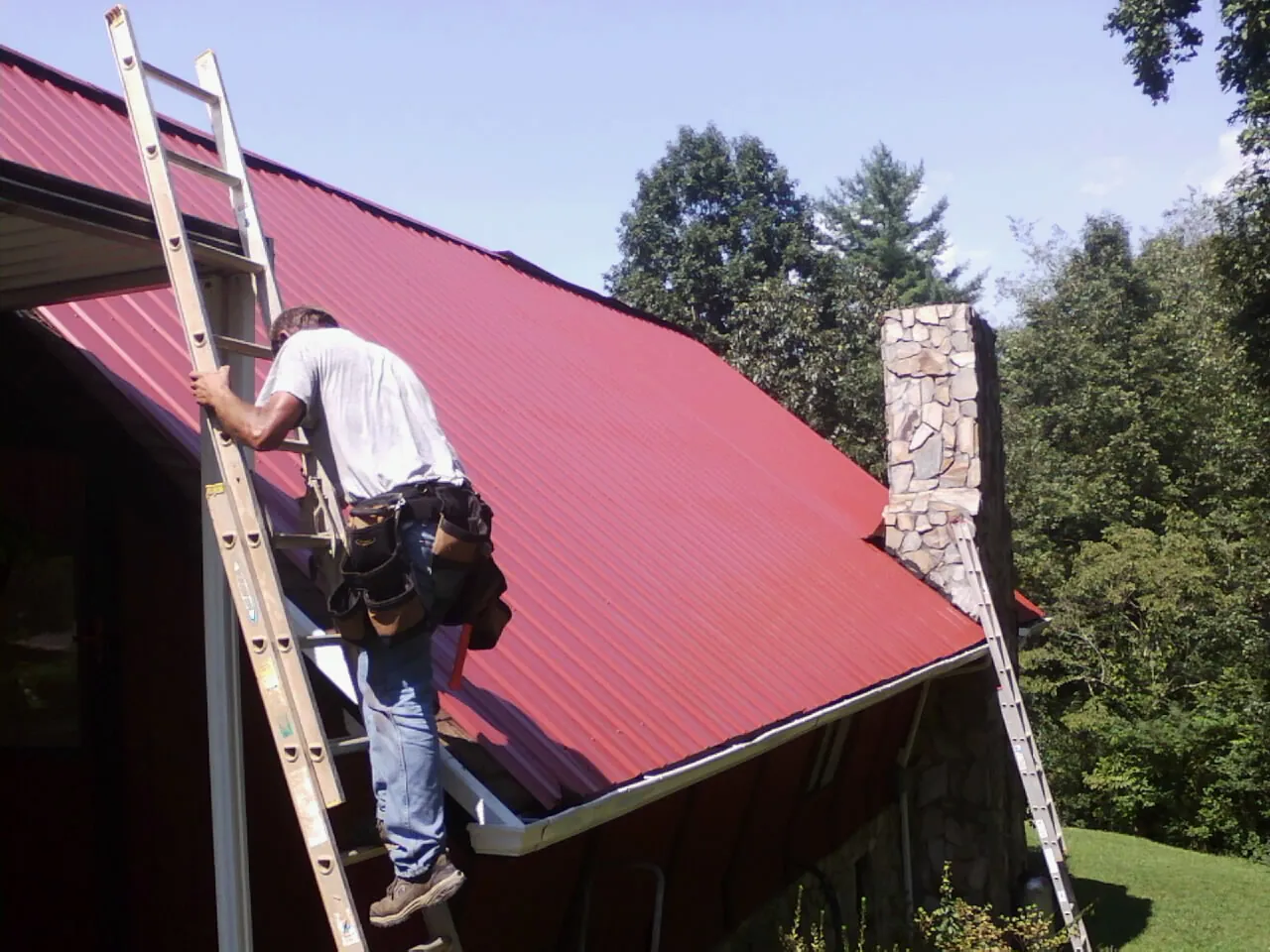Roofer on a ladder beside a red metal roof and stone chimney — Soffit & Fascia Repair by Roofing Brothers in Kingwood
